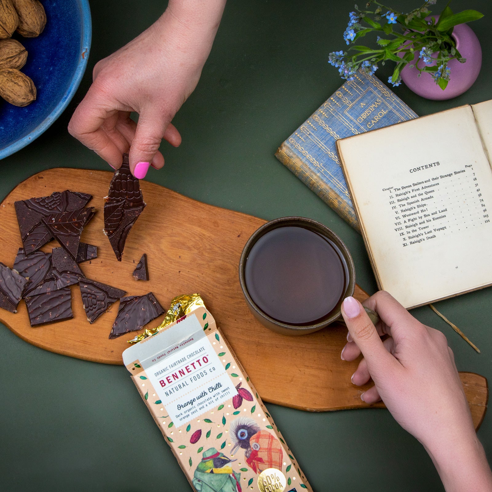 A bar of Bennetto Natural Foods organic, vegan and Fairtrade dark chocolate, Orange with Chilli flavour, lying down and open next to a cup of tea with two hands sharing