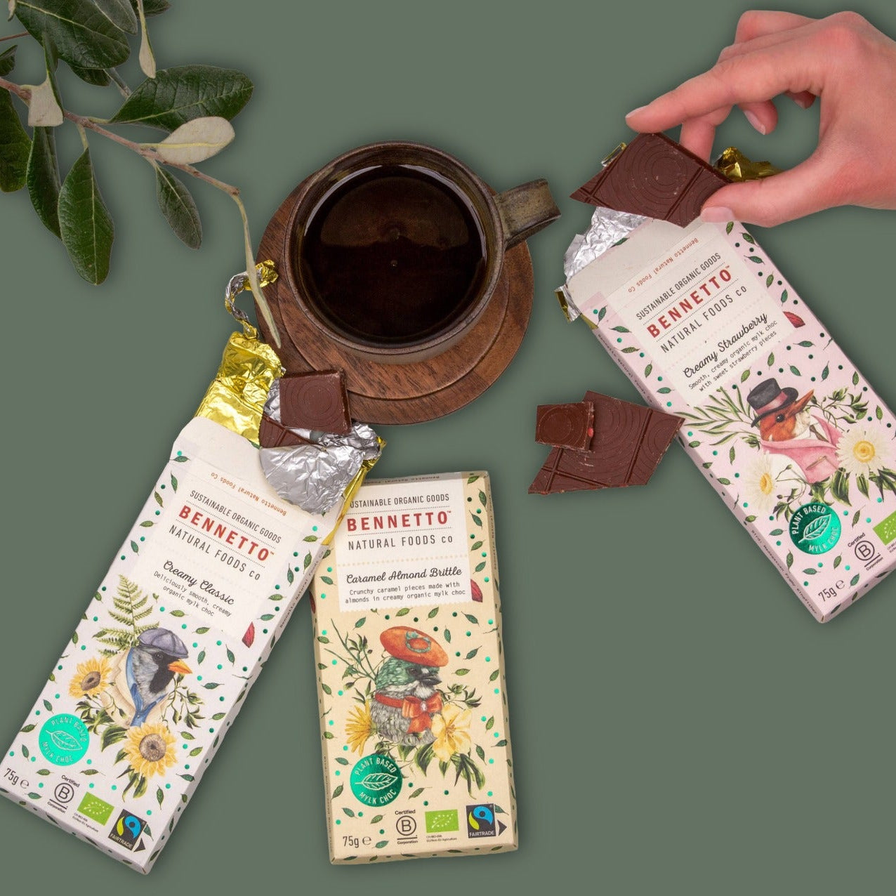 Three bars of Bennetto chocolate with illustrated pastel-colored wrappers are arranged on a muted green background alongside a dark ceramic cup and a hand holding a piece of chocolate. The wrappers feature botanical illustrations and text including 'BENNETTO' and 'Natural Food Co'.