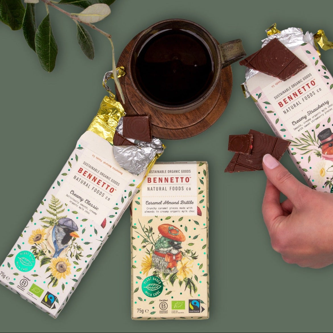 Three Bennetto chocolate bars with illustrated wrappers are arranged on a muted green background, alongside a dark cup of coffee, a small plant, and a hand holding a broken piece of chocolate.