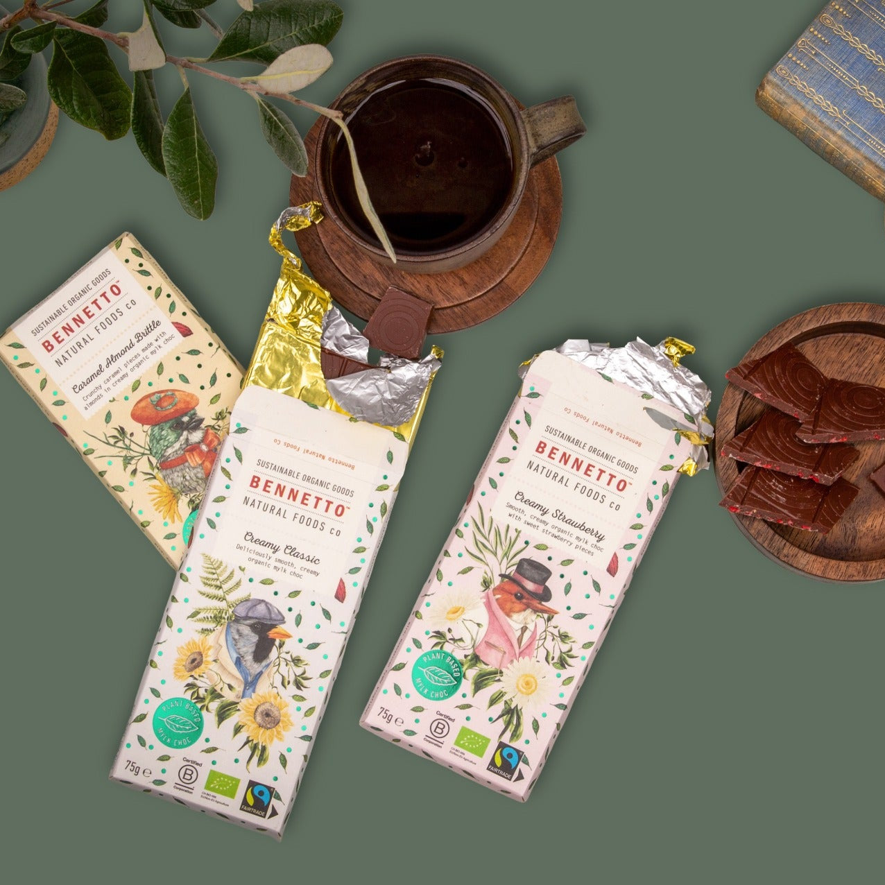 Two unwrapped Bennetto chocolate bars and one wrapped bar from the brand, arranged in a styled flat-lay with a ceramic mug, a book, and a wooden dish on a muted green background.