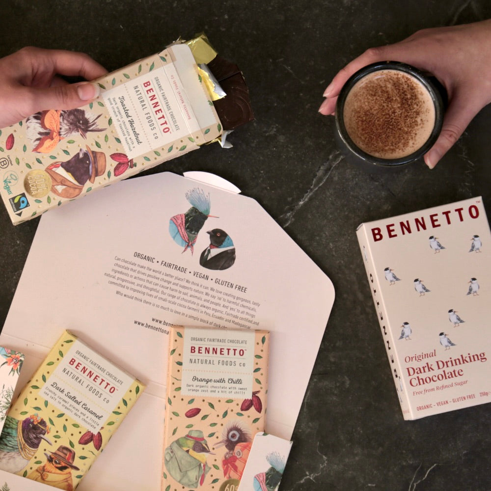 Lifestyle flatlay of 3 Bennetto chocolate bars and box of drinking chocolate. One hand holding ready to drink hot chocolate while other hand is holding open chocolate bar. 