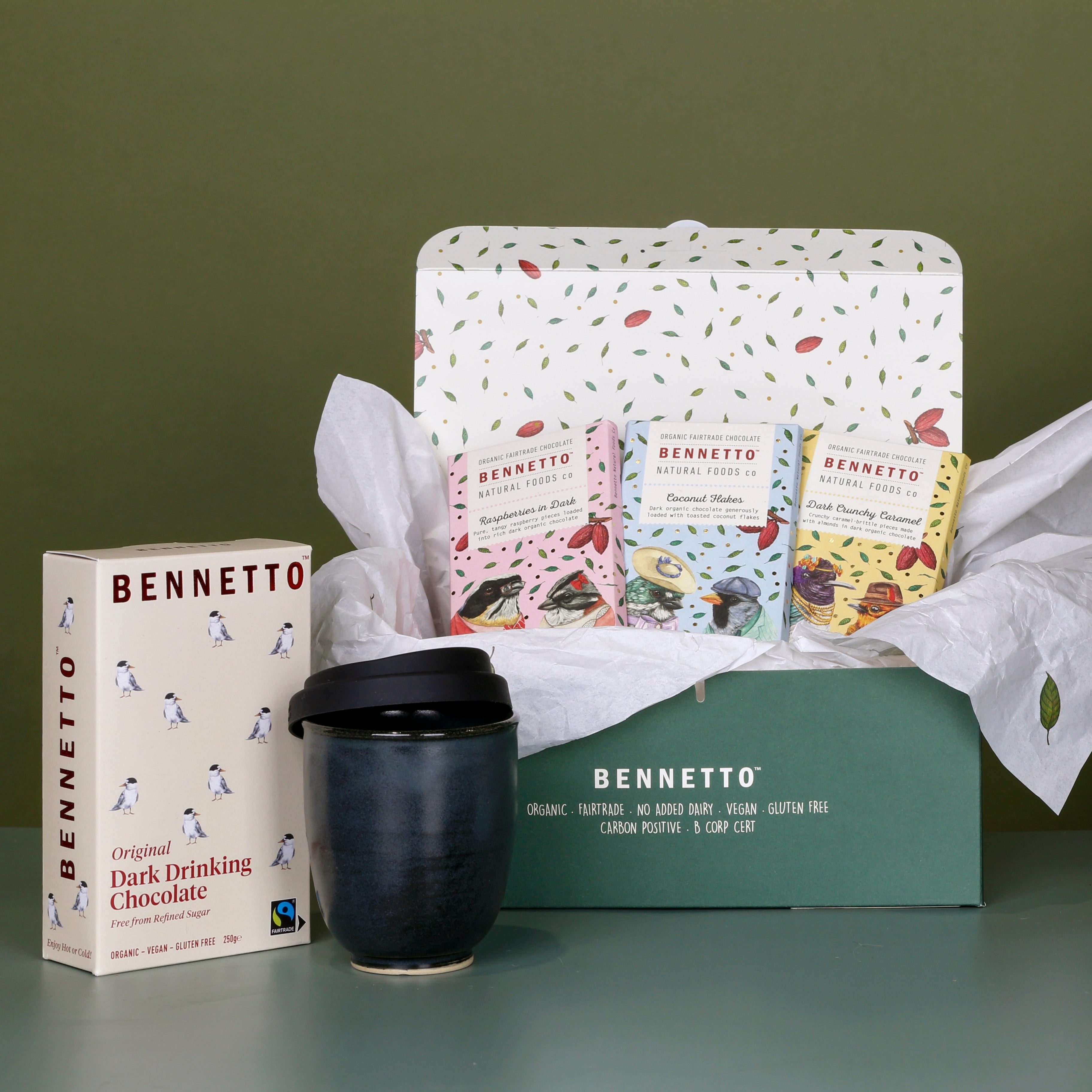 3 x Bennetto chocolate bars: Raspberries in Dark, Coconut Flakes, Dark Salted Caramel. In a green carton packaging, with tissue paper, a Bennetto Dark drinking Chocolate, with a handcrafted ceramic travel mug from the West Coast, on a green background.