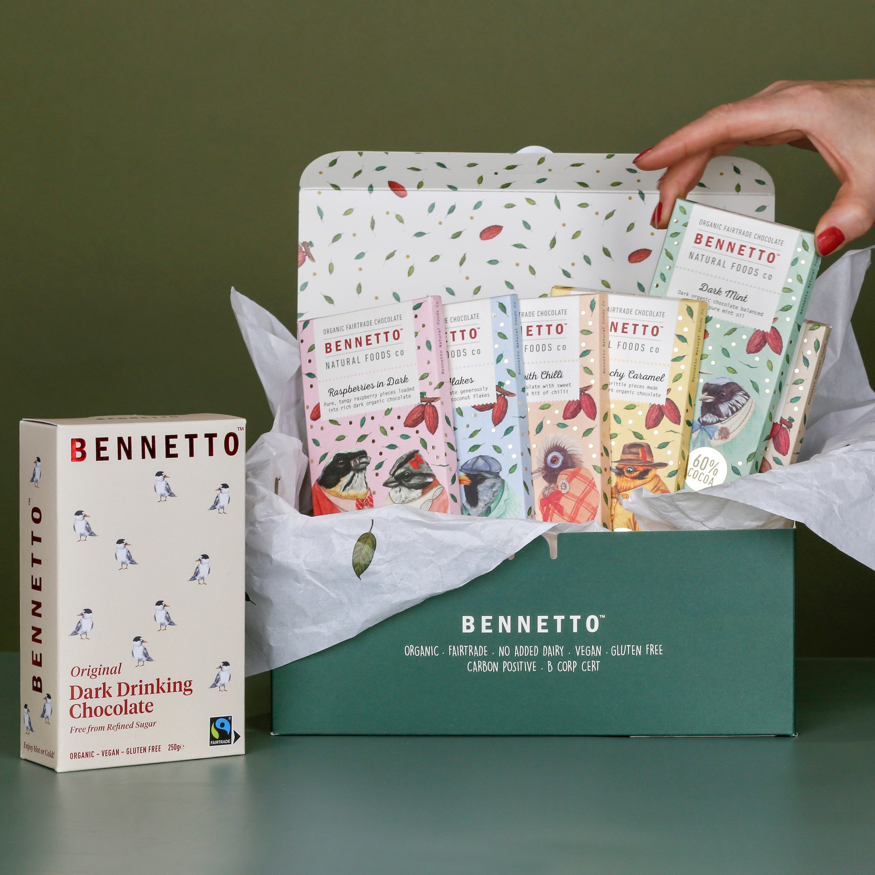Gift box with 6 x 80g Bennetto Chocolate bars: Raspberries in Dark, Coconut Flakes, Orange with Chilli, Dark Salted Caramel, Dark Mint, Toasted Hazelnut with a 250g Original Dark Chocolate. All in a dark green box with tissue paper. A hand reaching in picking up the Dark Mint.
