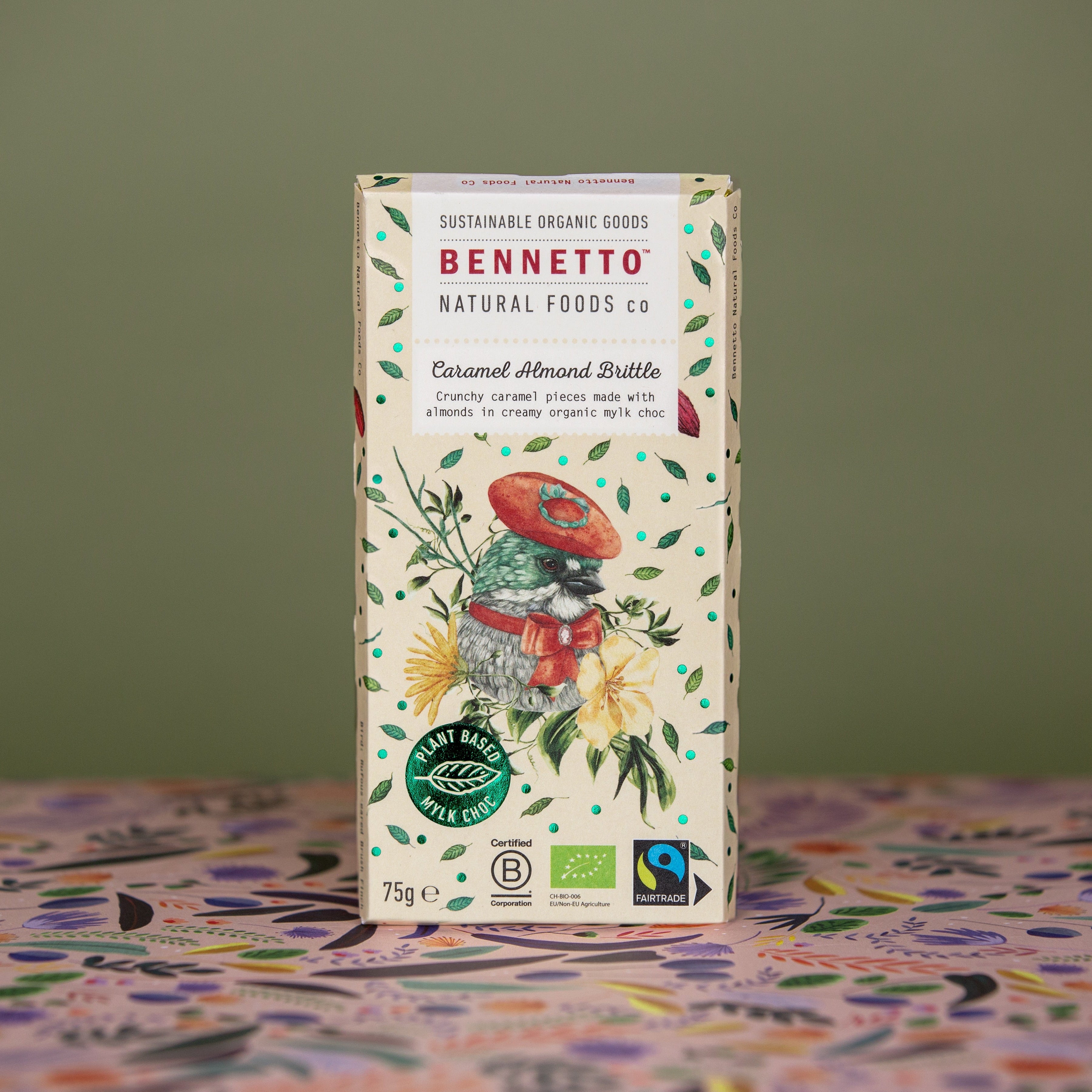 Bennetto Caramel Almond Brittle Mylk Chocolate standing upright on a green khaki background with patterned floral paper on the bottom. A 75g bar with bcorp, organic and fairtrade certifications. A light yellow with a bird and florals on the front packaging.