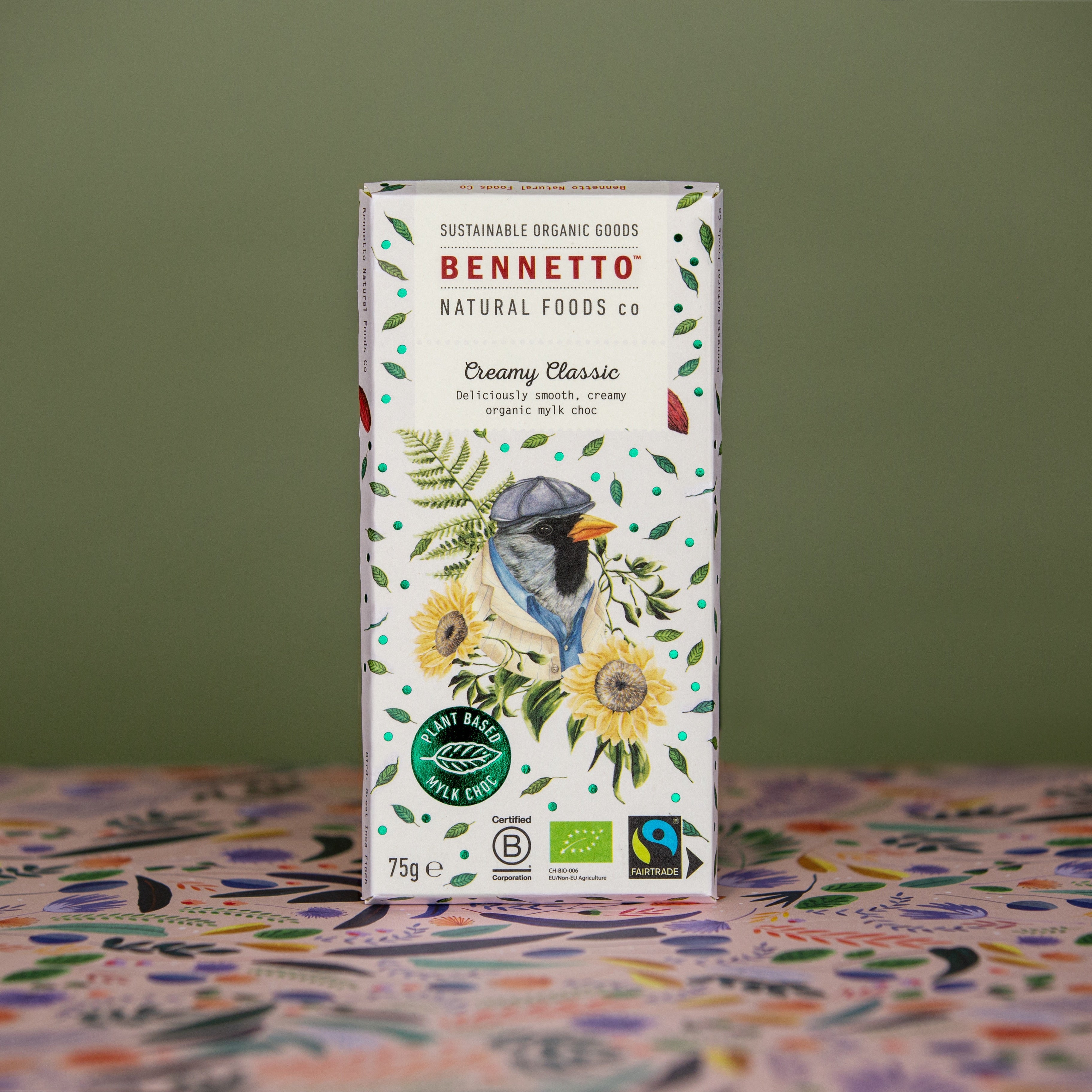 Bennetto Creamy Classic Mylk Chocolate standing upright on a green khaki background with patterned floral paper on the bottom. A 75g bar with bcorp, organic and fairtrade certifications. A light blue  with a bird and florals on the front packaging.