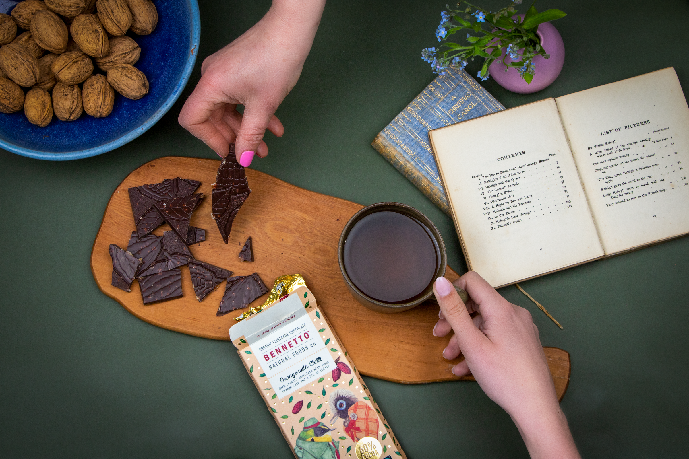Person holding a cup of tea next to a plate of Bennetto Natural Foods Orange with. Chilli chocolate, a book, and a bowl of walnuts on a green surface.